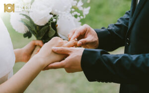 Close-up of one person placing a ring on another’s finger during a wedding. The person receiving the ring holds a bouquet.