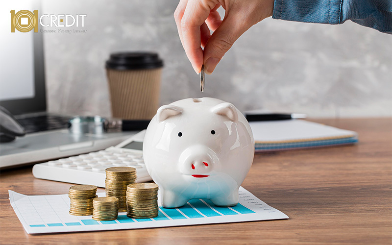 Hand placing a coin into a white piggy bank. Stacks of coins, calculator, and desk items suggest saving and budgeting