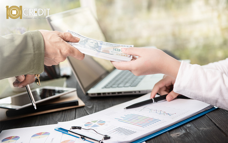 One person handing a stack of money to another across a desk with financial charts and a laptop.