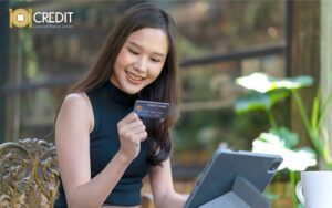 Person outdoors holding a credit card and using a tablet. Smiling, with plants and a coffee mug nearby