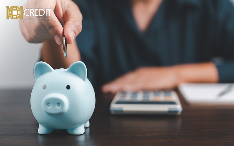 Person placing a coin into a light blue piggy bank. Calculator and notebook on the table suggest saving and budgeting.