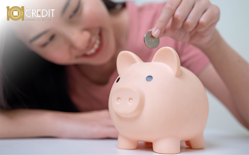 Smiling person holding a coin above a light pink piggy bank. White surface and softly blurred background suggest saving and positivity.