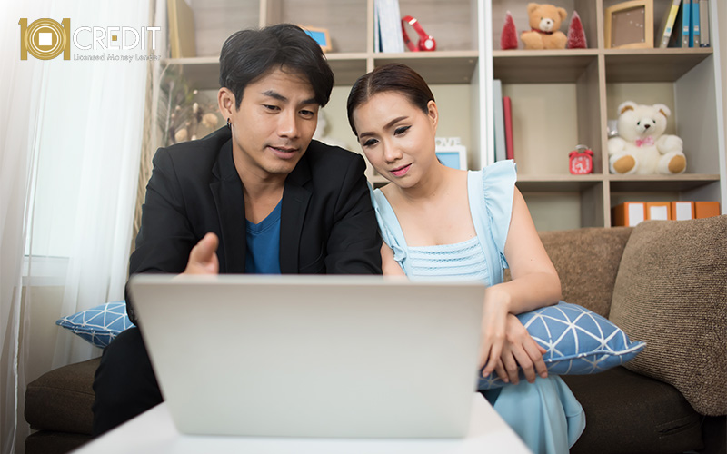 Two people sitting on a couch, focused on a laptop screen, with a bookshelf in the background.