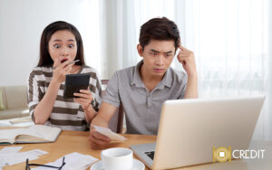 A shocked woman looking at a calculator while a stressed man reviews financial documents
