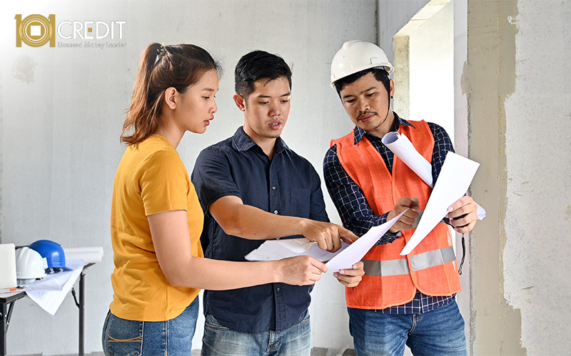 Three people reviewing documents at a construction site—one in a white helmet and orange vest, others in casual clothing—surrounded by building materials.