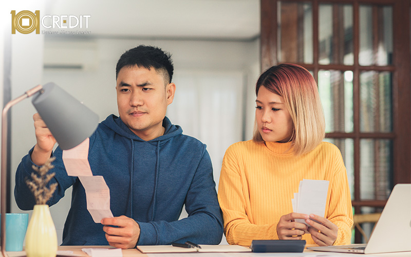 Two people reviewing receipts and financial documents at a home table with a calculator, laptop, and notebook—one in a blue hoodie, the other in a yellow sweater.
