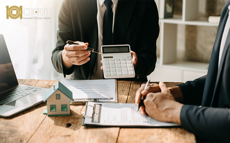 Two professionals in business attire discussing financial documents at a table with a house model, calculator, and laptop—suggesting a mortgage or loan consultation.