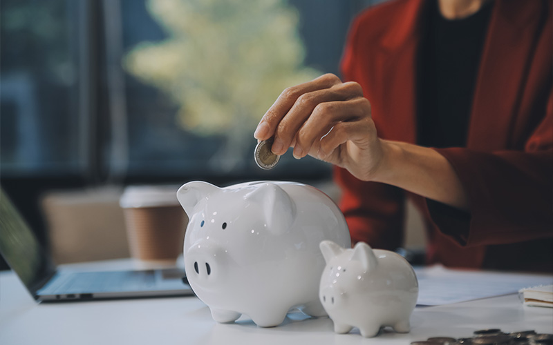 A close-up of a person in a red blazer dropping a coin into a white piggy bank