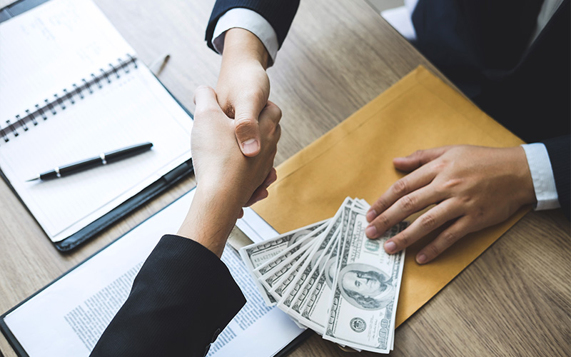 Two professionals shaking hands over a desk with cash and documents